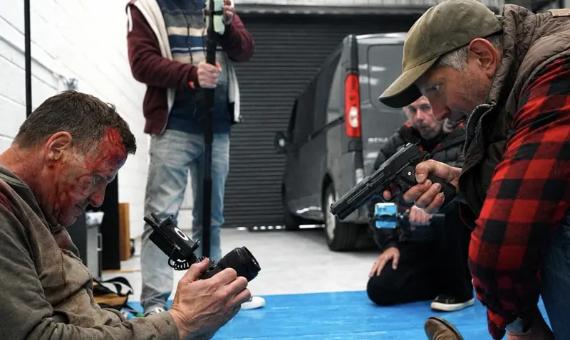 Andy Blithe directing an action scene on set, camera on a bloodied actor and a man in a cap aiming a gun