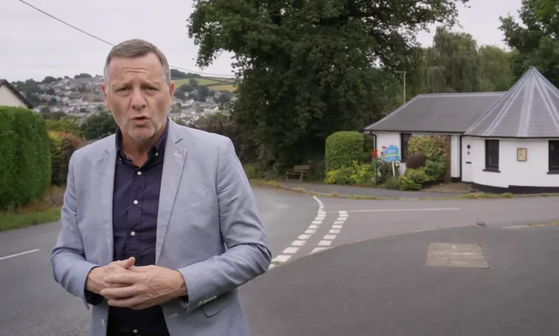 Andy Blithe presenting to camera in a grey blazer by a country road
